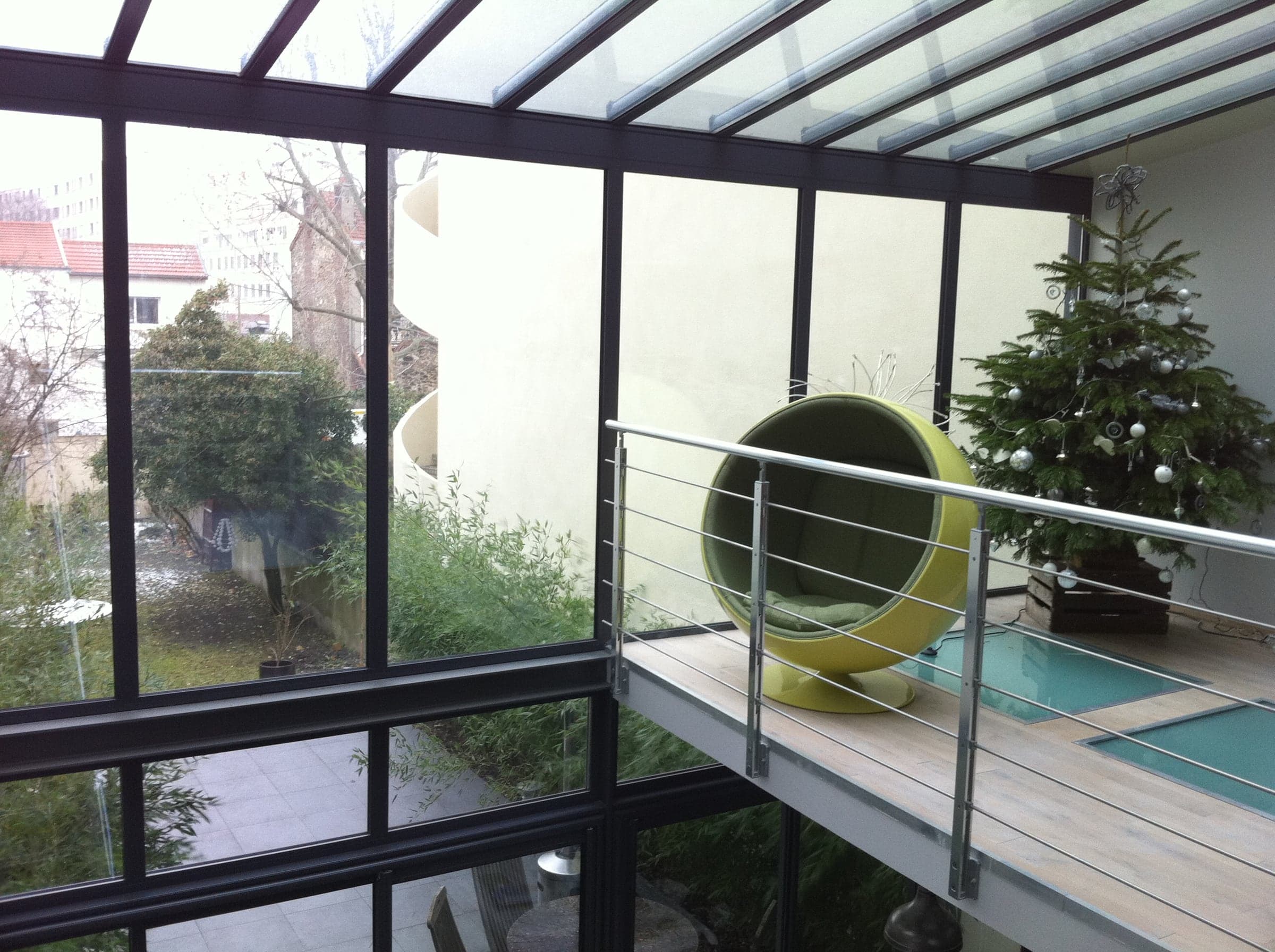 Véranda contemporaine à structure noire, passerelle vitrée et fauteuil boule au-dessus d’un patio