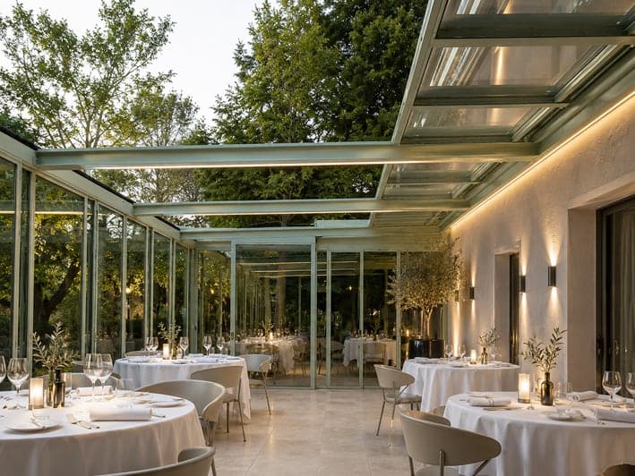 Véranda de restaurant à toiture vitrée ouvrante, tables dressées le soir sous la verrière